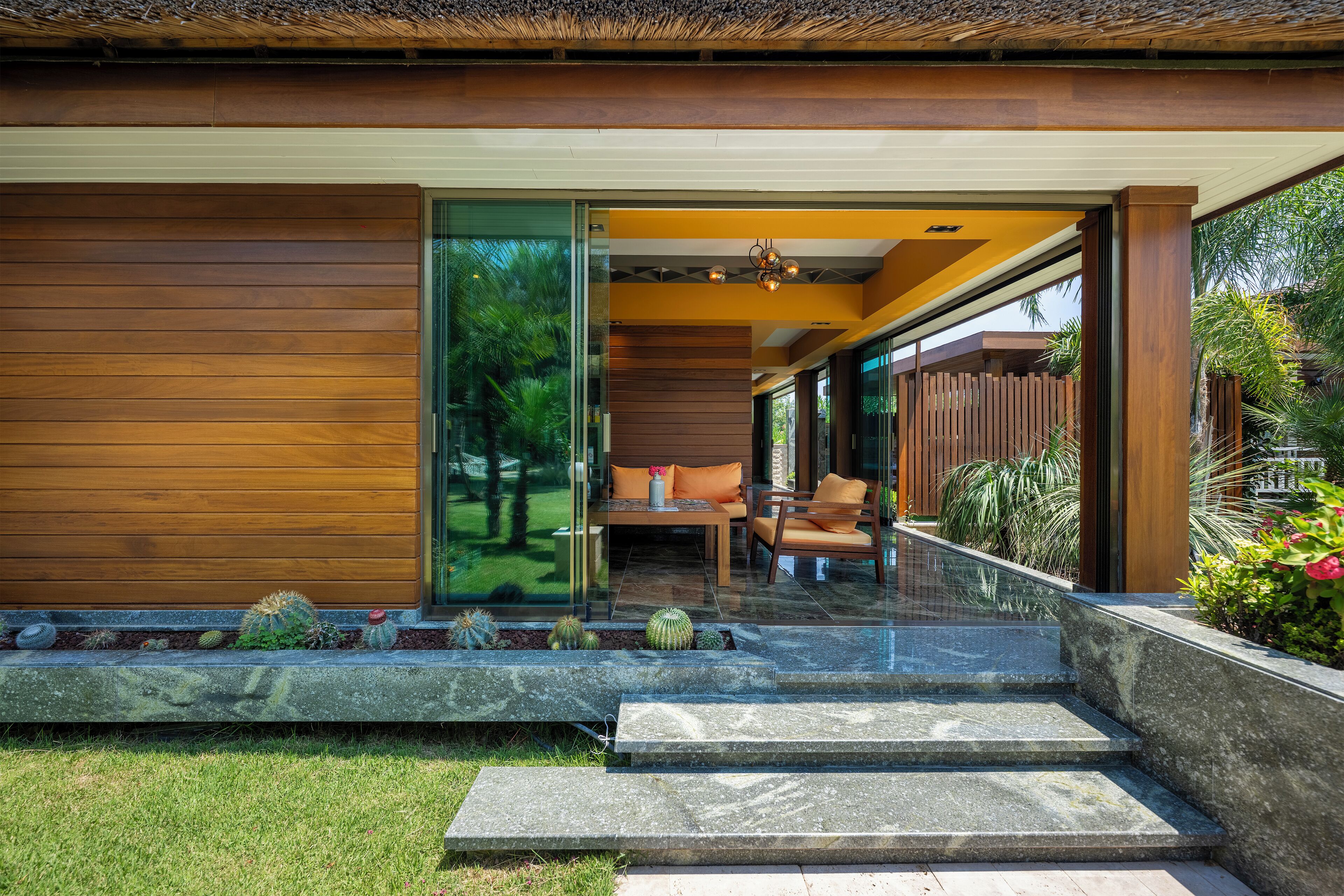 A modern house with a wooden exterior, a glass door leading to an outdoor seating area with a table and chairs, and a garden with various plants and a stone pathway.