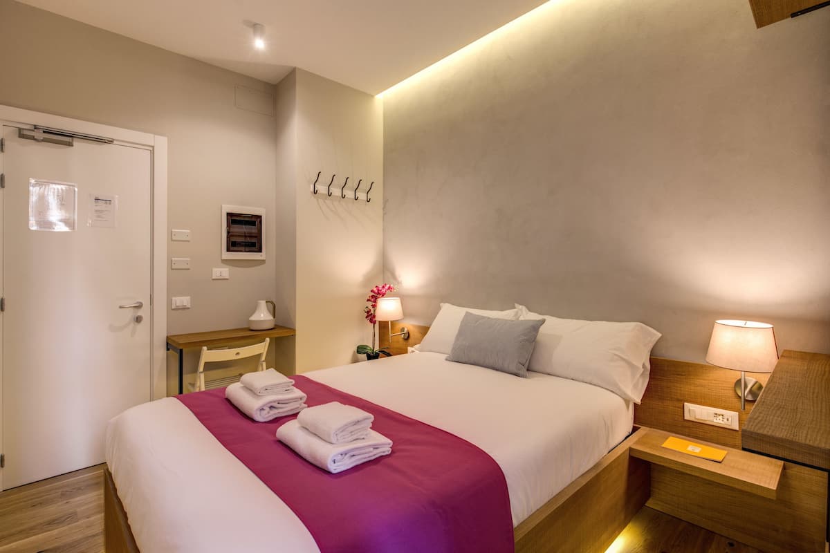 A hotel room with a neatly made bed, a bedside table with a lamp, a wall-mounted light, and a door with a sign.
