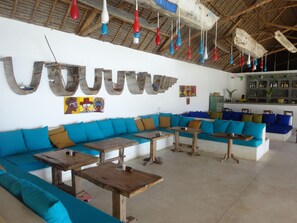 A spacious lounge area with a large wooden sofa, multiple tables, and hanging light fixtures. - Nur Beach Hotel (Jambiani)