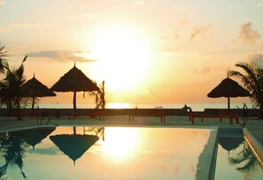 A pool with a sunset view, palm trees, and beach umbrellas.