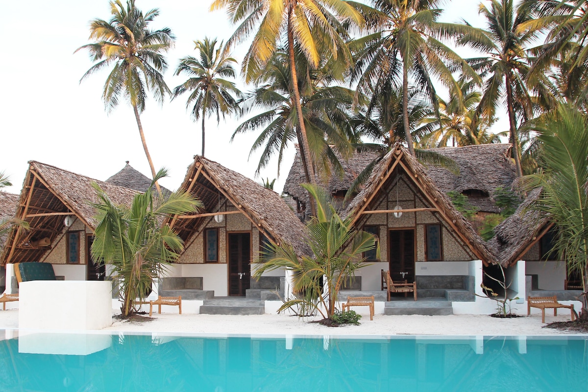 A poolside area with thatched-roof huts, palm trees, and a clear blue pool.