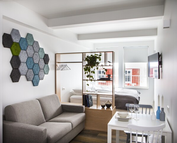 A modern, well-lit room with a grey sofa, a white table with plates and glasses, a TV, and a wall-mounted hexagonal art piece. - Island Apartments (Reykjavik)