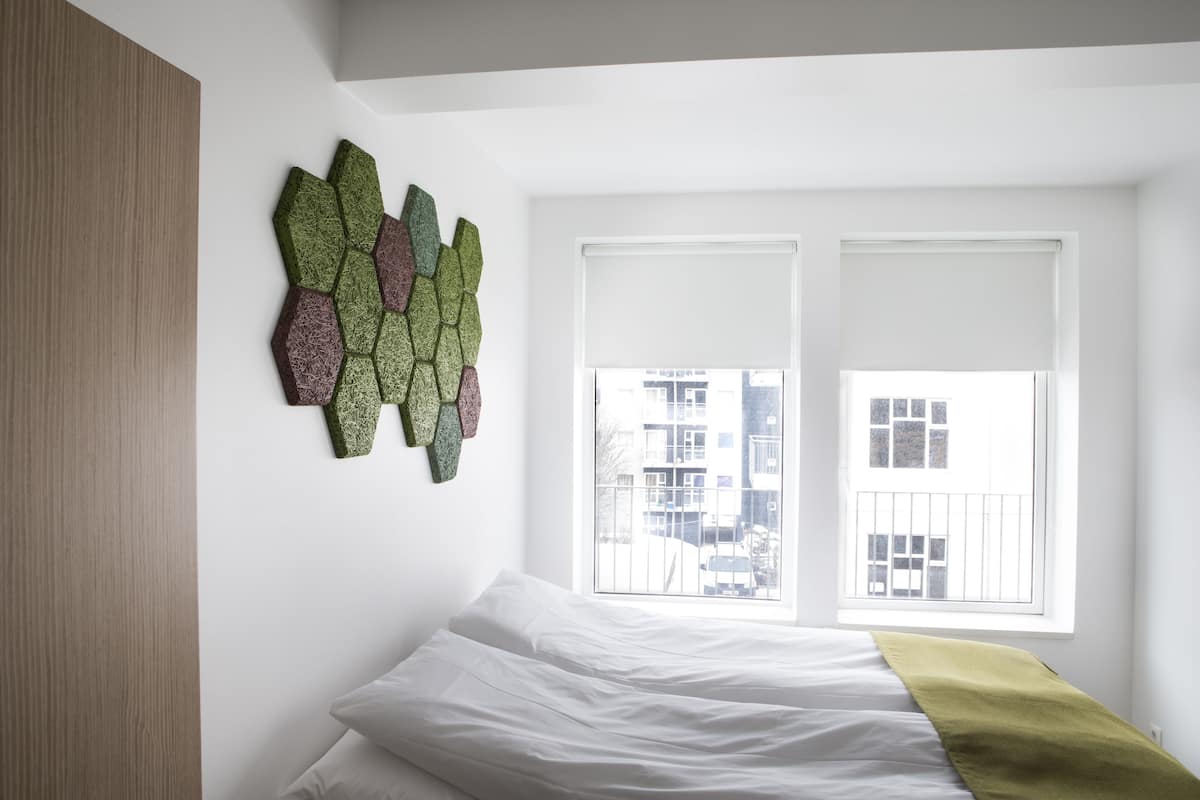 Une chambre à coucher avec un lit, des murs blancs, une fenêtre avec des stores et une œuvre d’art géométrique accrochée au mur.