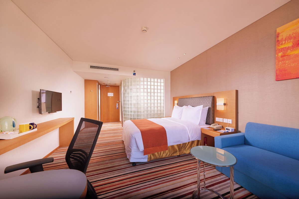 A hotel room with a bed, a desk, a chair, a sofa, a TV, and a window with blinds.