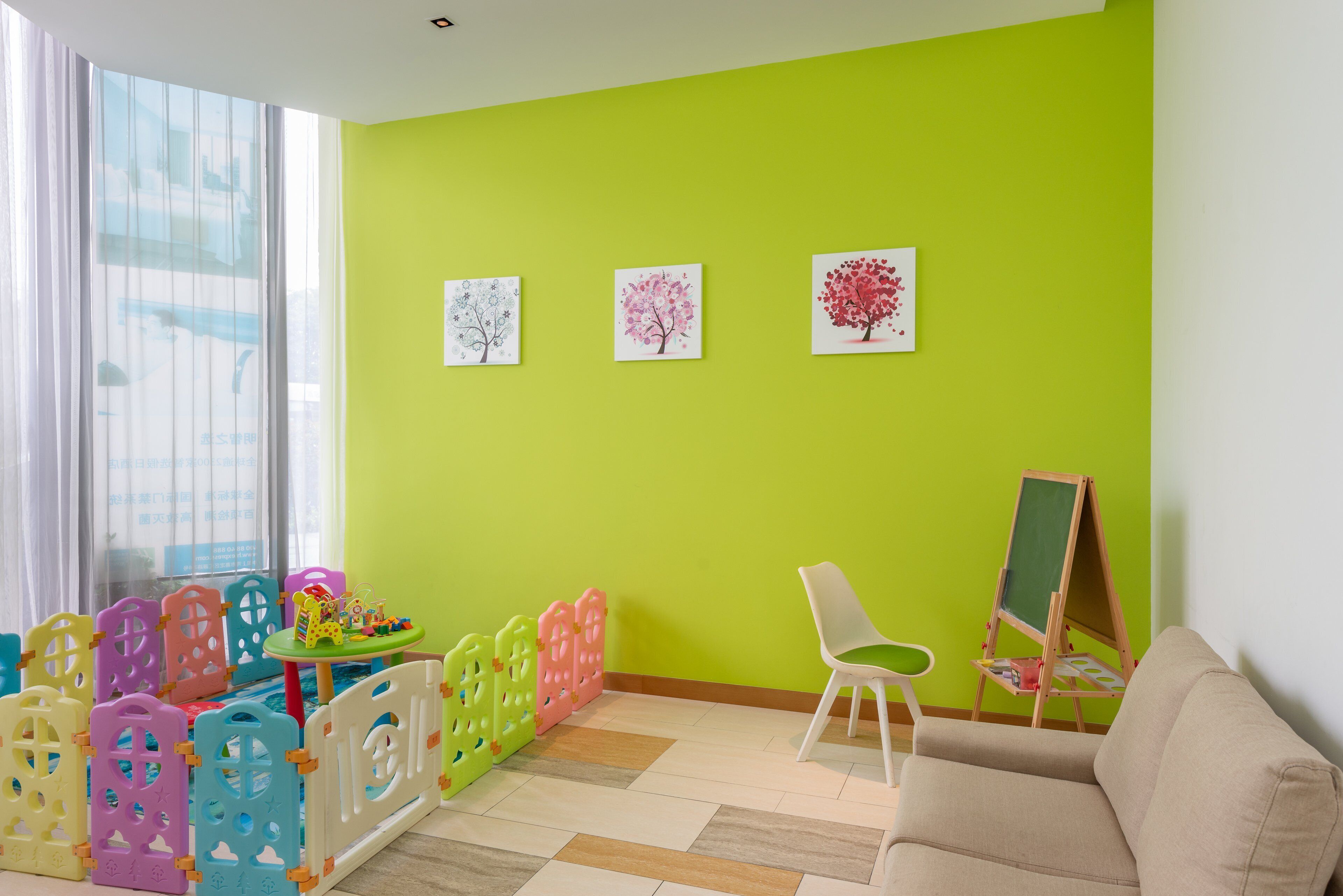 Une pièce avec un mur vert, une fenêtre avec des stores, une chaise blanche, une petite table avec des jouets et un tableau noir.