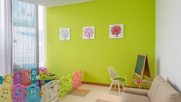 Une pièce avec un mur vert, une fenêtre avec des stores, une chaise blanche, une petite table avec des jouets et un tableau noir.