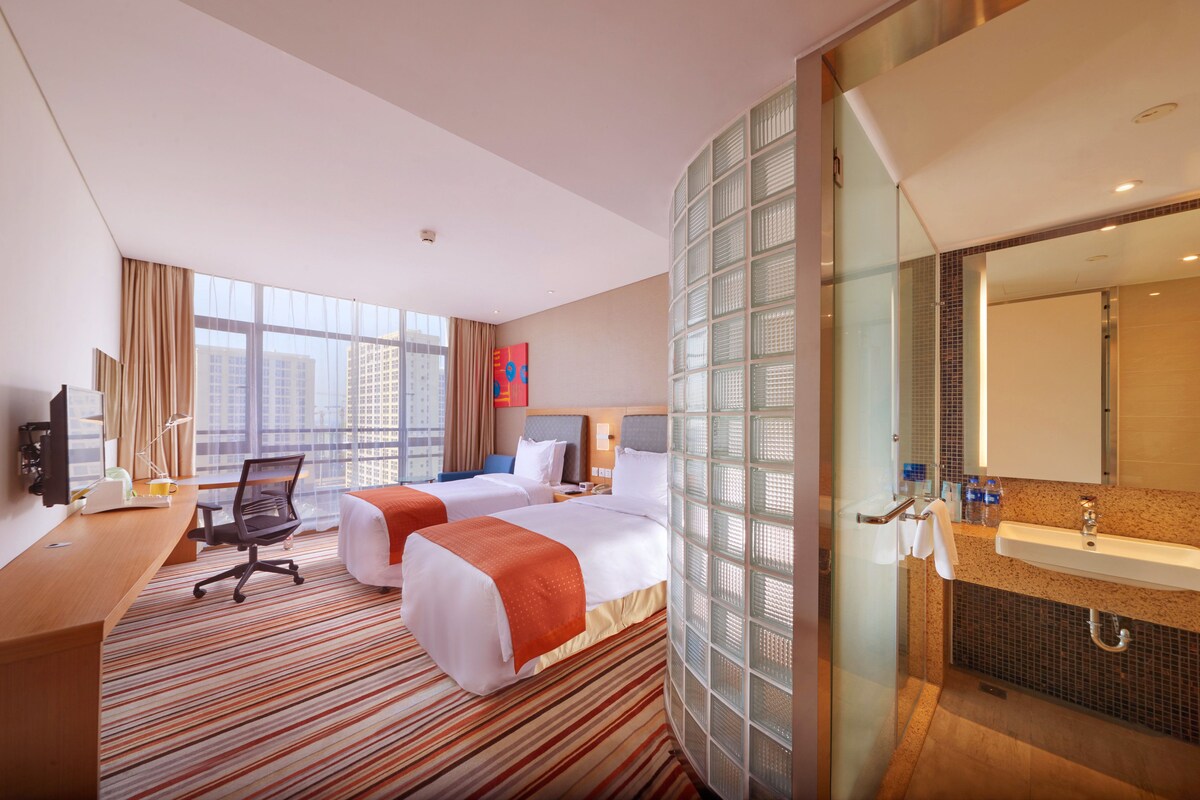 Quarto de hotel com duas camas, uma escrivaninha, uma cadeira, uma janela grande com vista para edifícios e banheiro com pia e espelho.