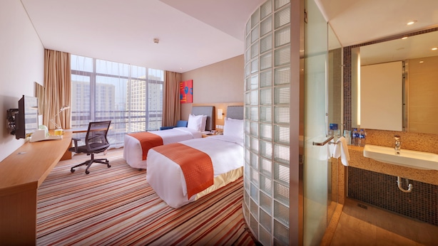 A hotel room with two beds, a desk, a chair, a large window with a view of buildings, and a bathroom with a sink and mirror. - Holiday Inn Express Shanghai Jiading Industry Park by IHG (Shanghai)