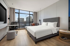 Une chambre d’hôtel moderne, dotée d’un grand lit, d’un bureau, d’une chaise et offrant une vue sur le paysage urbain grâce à de grandes fenêtres.