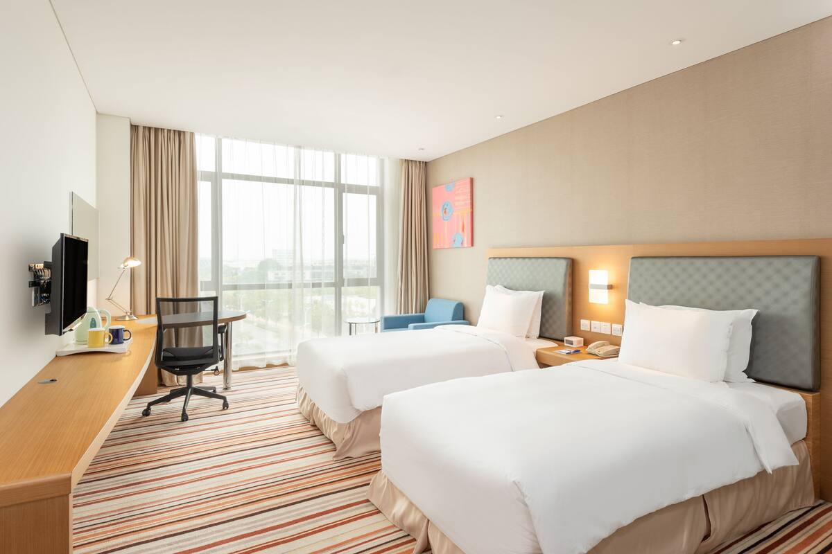 A hotel room with two beds, a desk, a chair, a TV, and a window with curtains.
