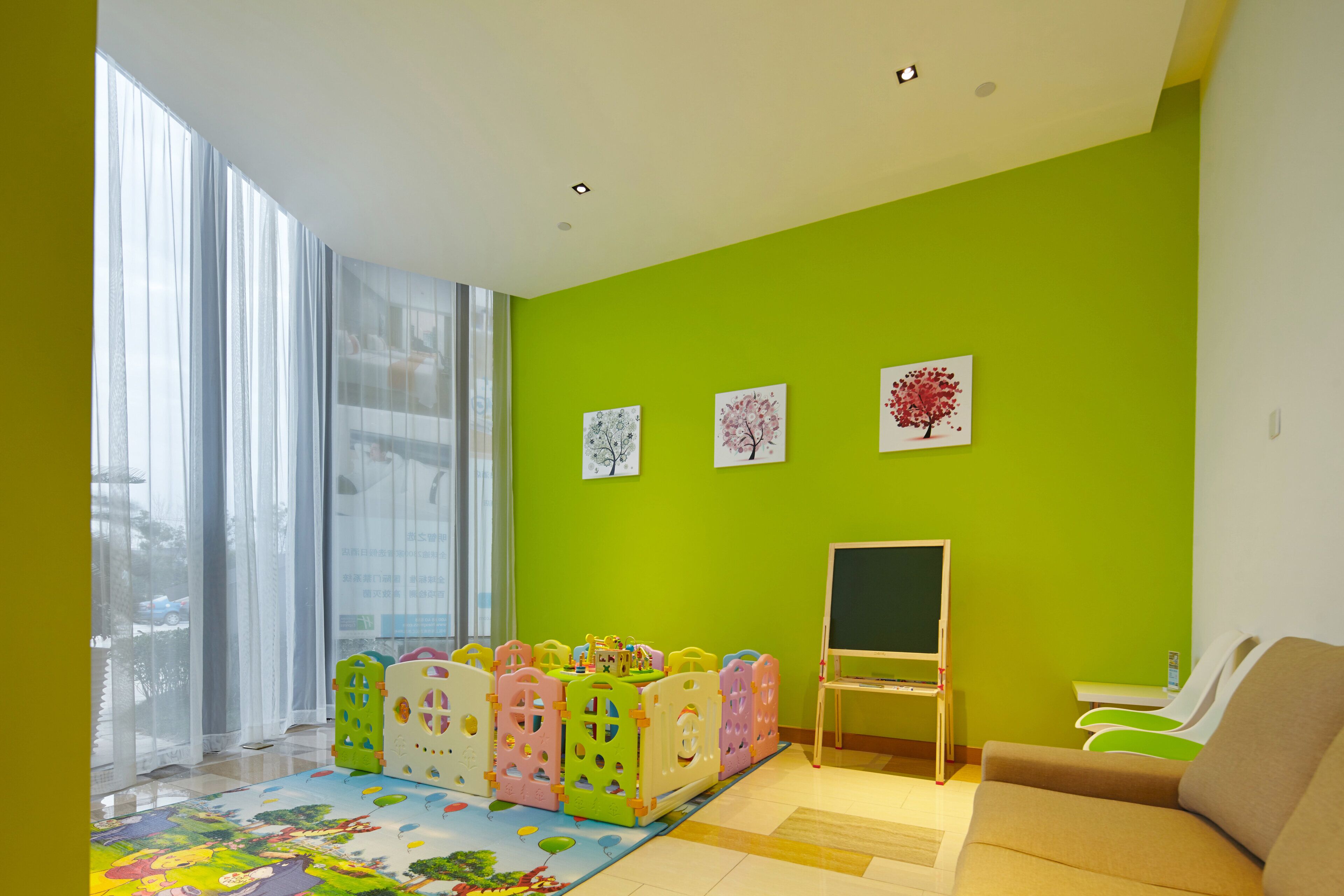 A bright room with a green wall, a play area with colorful playpens, a blackboard, and a sofa.