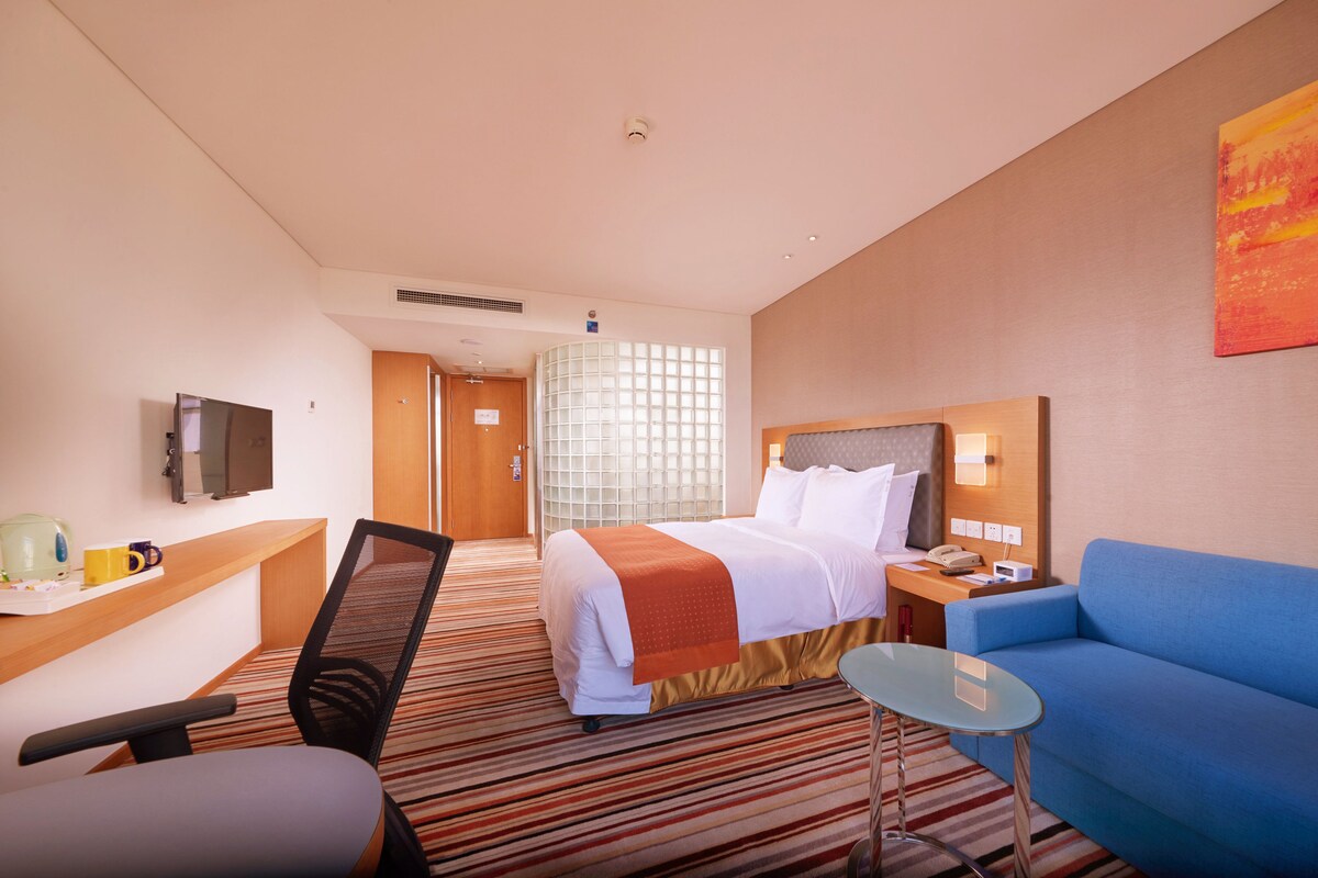 Quarto de hotel com cama, secretária com computador, cadeira, sofá azul e uma pequena mesa.