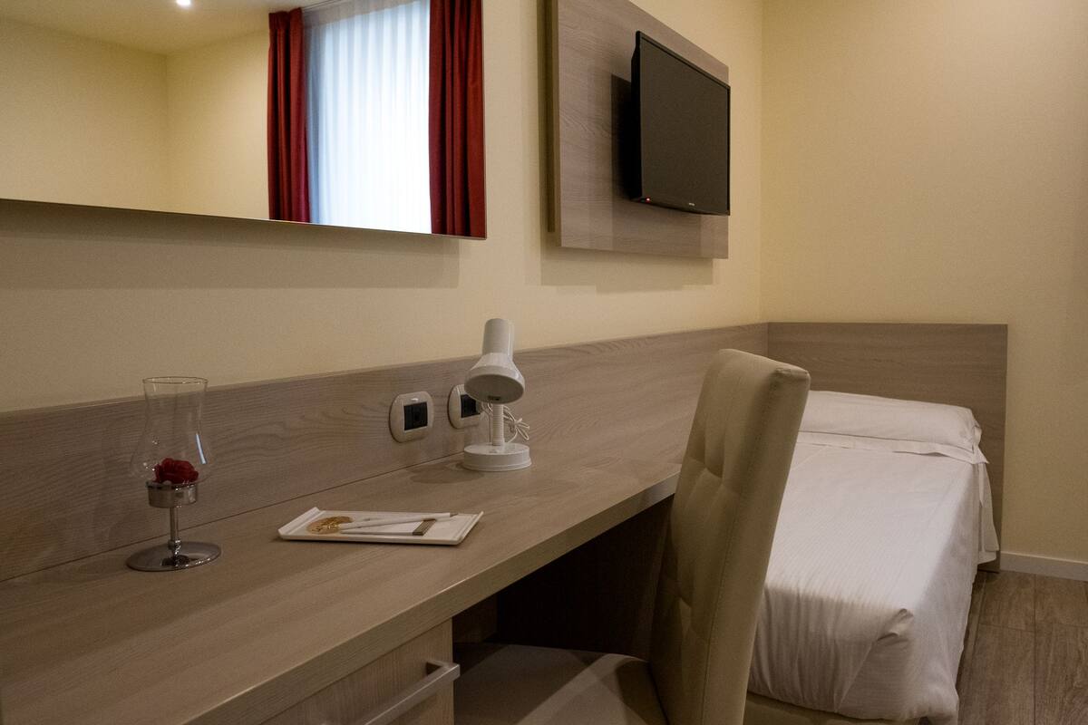 Een hotelkamer met een houten bureau, een flatscreen televisie aan de muur, een nachtkastje met een lamp, een stoel en een bed met witte lakens.