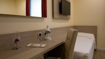 A hotel room with a wooden desk, a flat-screen TV mounted on the wall, a bedside table with a lamp, a chair, and a bed with white linens.