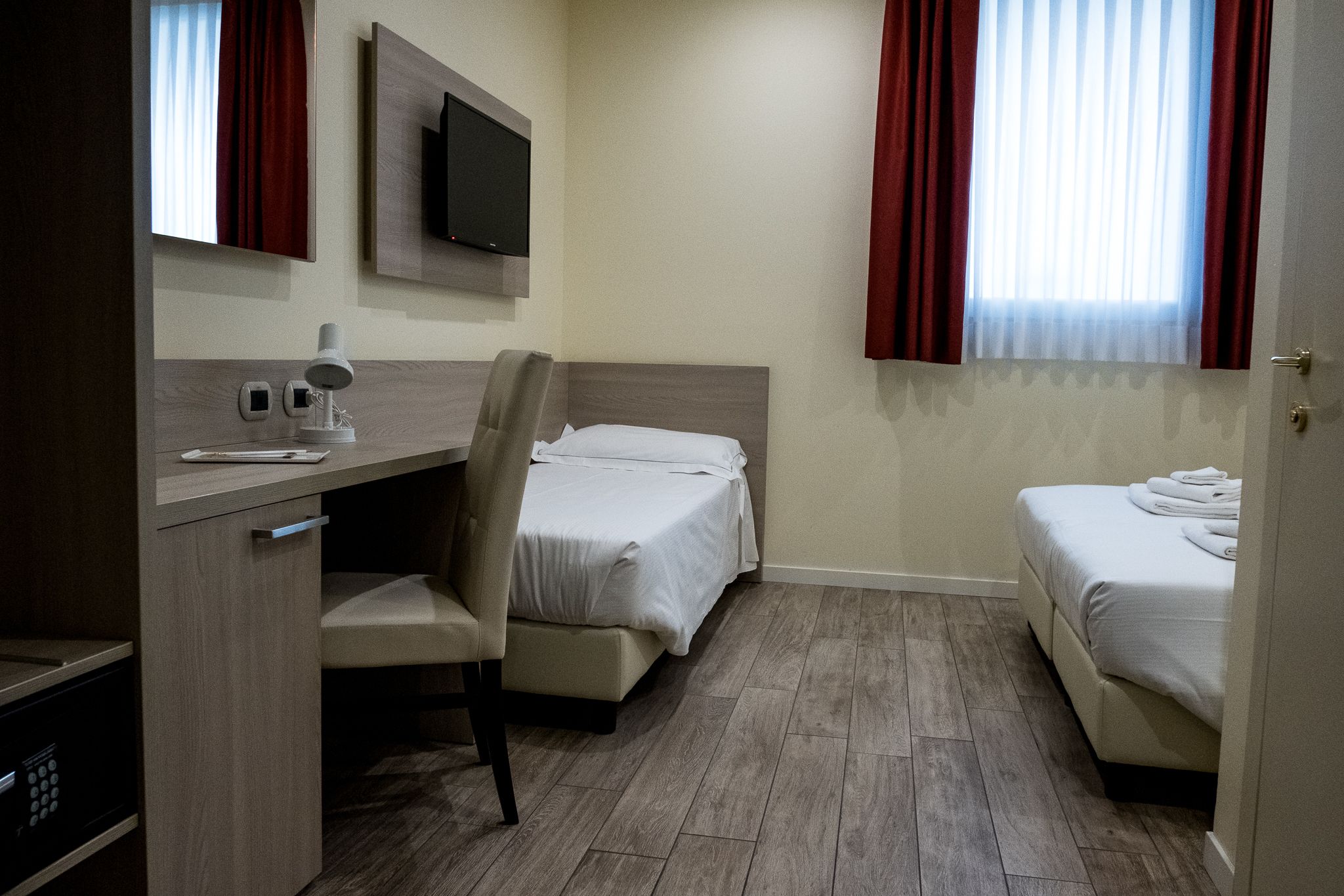 A hotel room with a desk, chair, two beds, a TV, and a window with curtains.