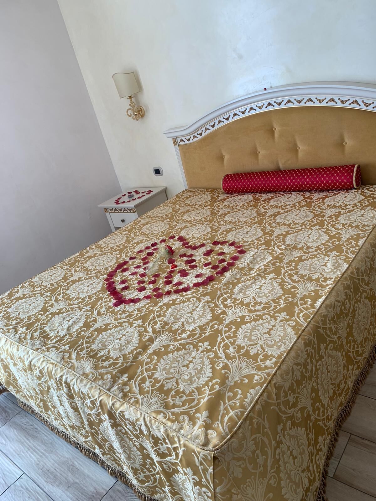 A neatly made bed with a patterned bedspread, a red pillow, and a wall-mounted lamp.