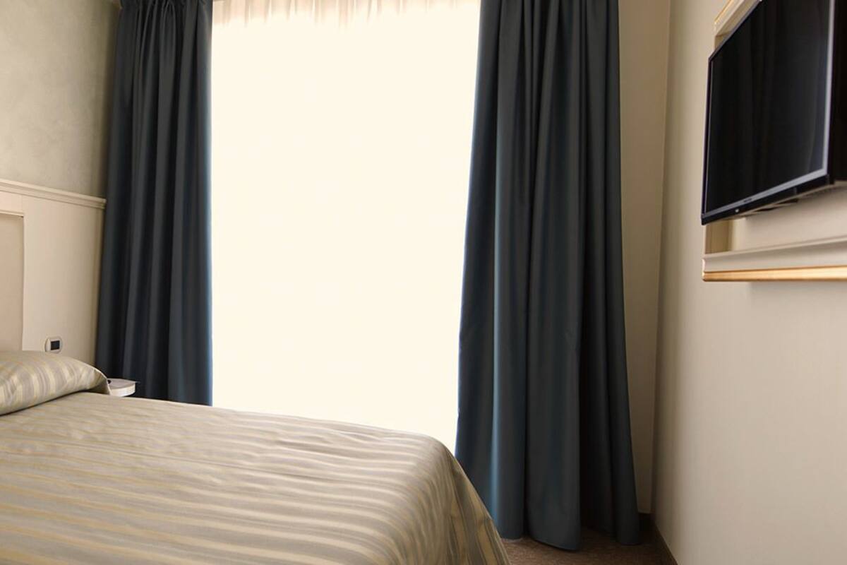 Hotelkamer met een bed, hoofdeinde, een aan de muur bevestigde televisie en een raam met gordijnen.