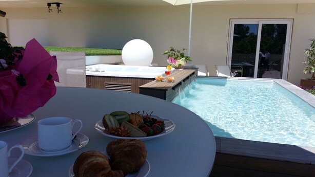 Un espace aménagé au bord de la piscine, avec un jacuzzi, une table à manger où sont servis des croissants et des fruits, et un grand parasol.