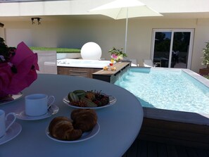 Een zwembadterras met een bubbelbad, een eettafel met croissants en fruit, en een grote parasol.