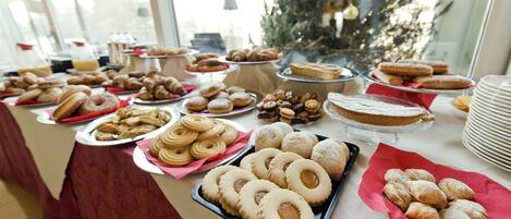 A buffet table with various pastries and desserts, a Christmas tree in the background, and a window view.