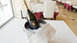 Une bouteille de champagne dans un seau en métal rempli de glace, posée sur une table dans une pièce où se trouvent des tables et des chaises blanches.