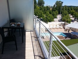 Un balcone con tavolo e sedie, una zona piscina con lettini e una vista su un parcheggio e alberi.