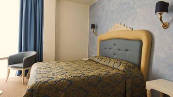 A hotel room with a bed, a chair, a small table, and a bottle of wine.