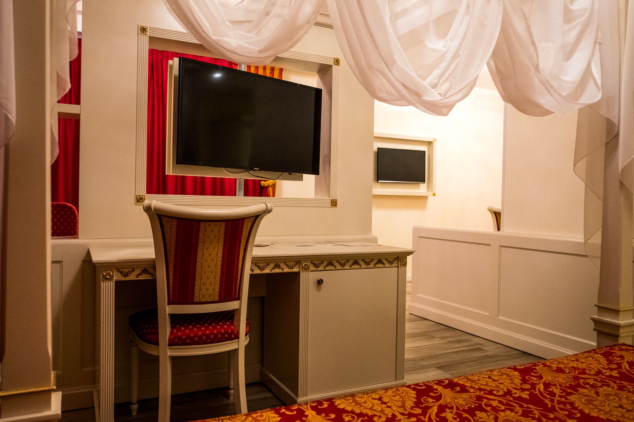 Una habitación con una televisión montada en la pared, un escritorio, una silla y una alfombra estampada.
