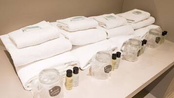 Des serviettes blanches, soigneusement pliées, sont empilées sur une étagère, à côté de flacons en verre transparent contenant des produits de toilette, étiquetés « Hygiène et sécurité ».