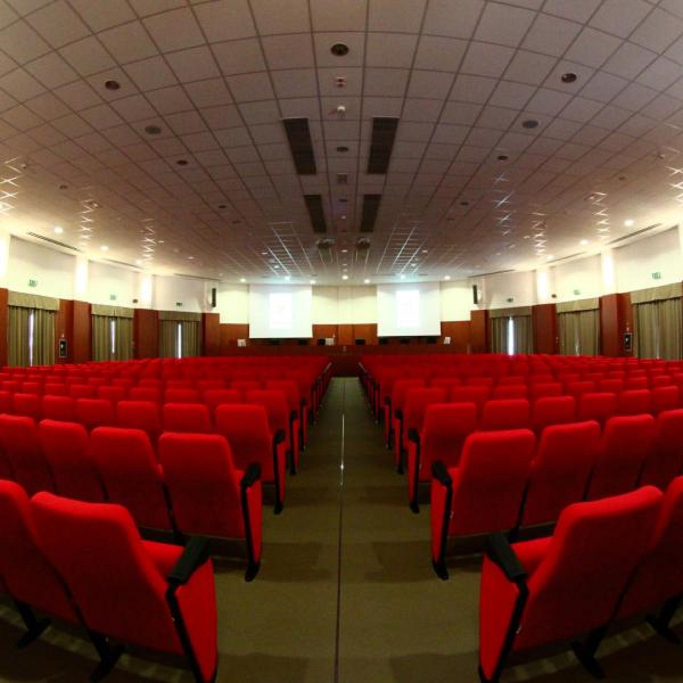 A conference room with rows of red chairs, a stage with a projector, and a grid-patterned ceiling.