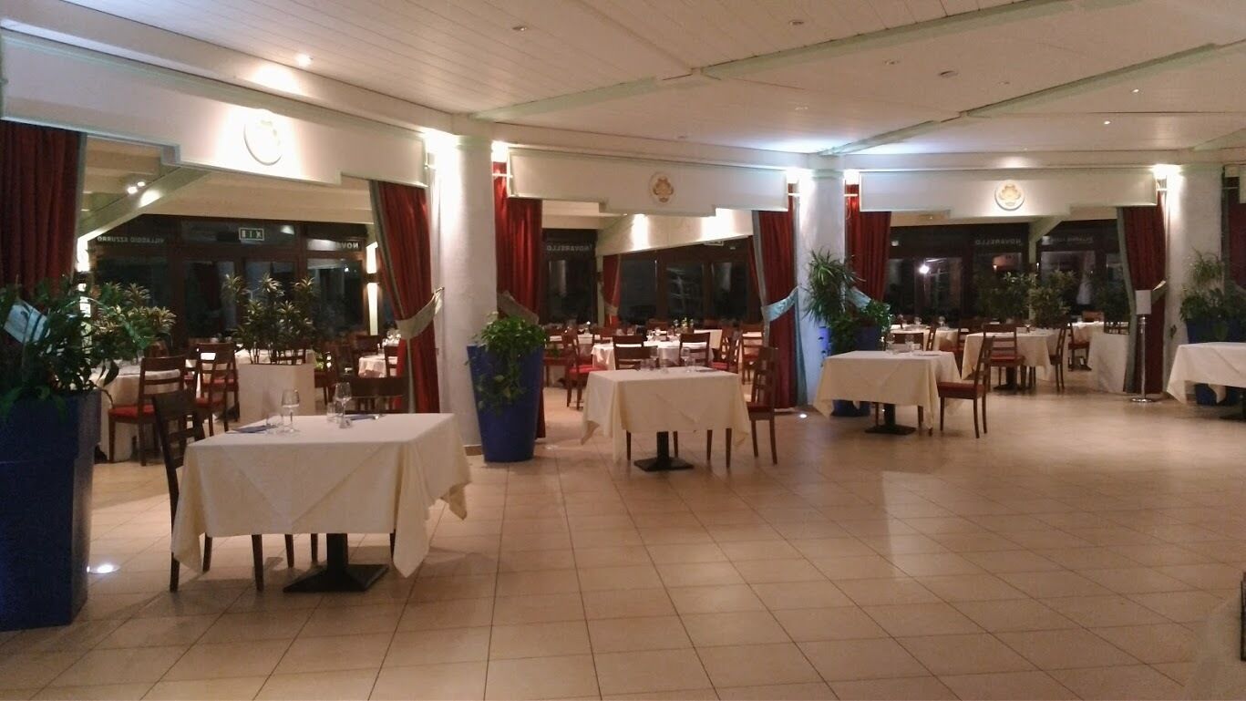 A spacious indoor dining area with multiple tables and chairs, large potted plants, and a tiled floor.