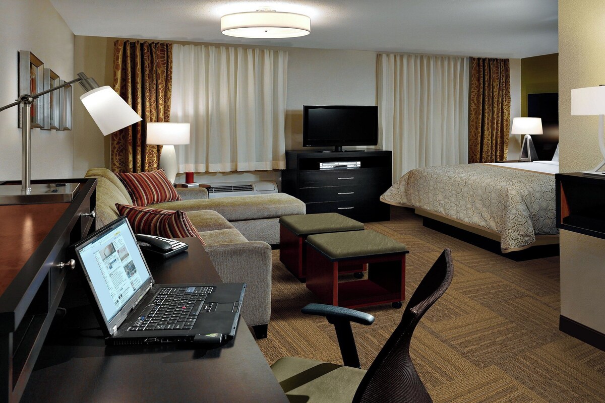 A hotel room with a bed, a desk with a laptop, a TV, and a bedside table.