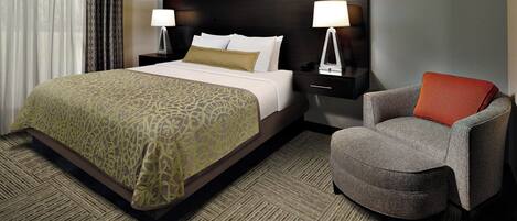 A hotel room with a large bed, two bedside tables with lamps, a grey armchair, and a patterned carpet.