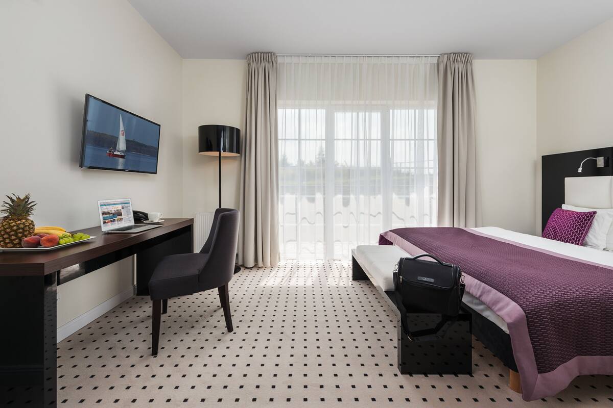 A hotel room with a bed, desk, chair, and a large window with curtains.