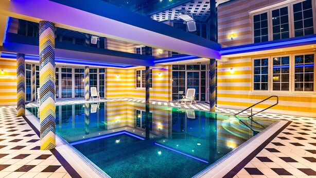 A modern indoor swimming pool with geometric patterned tiles, a central pool area, and seating areas with chairs. - Boutique Apartmenty M&M (Mikolajki)