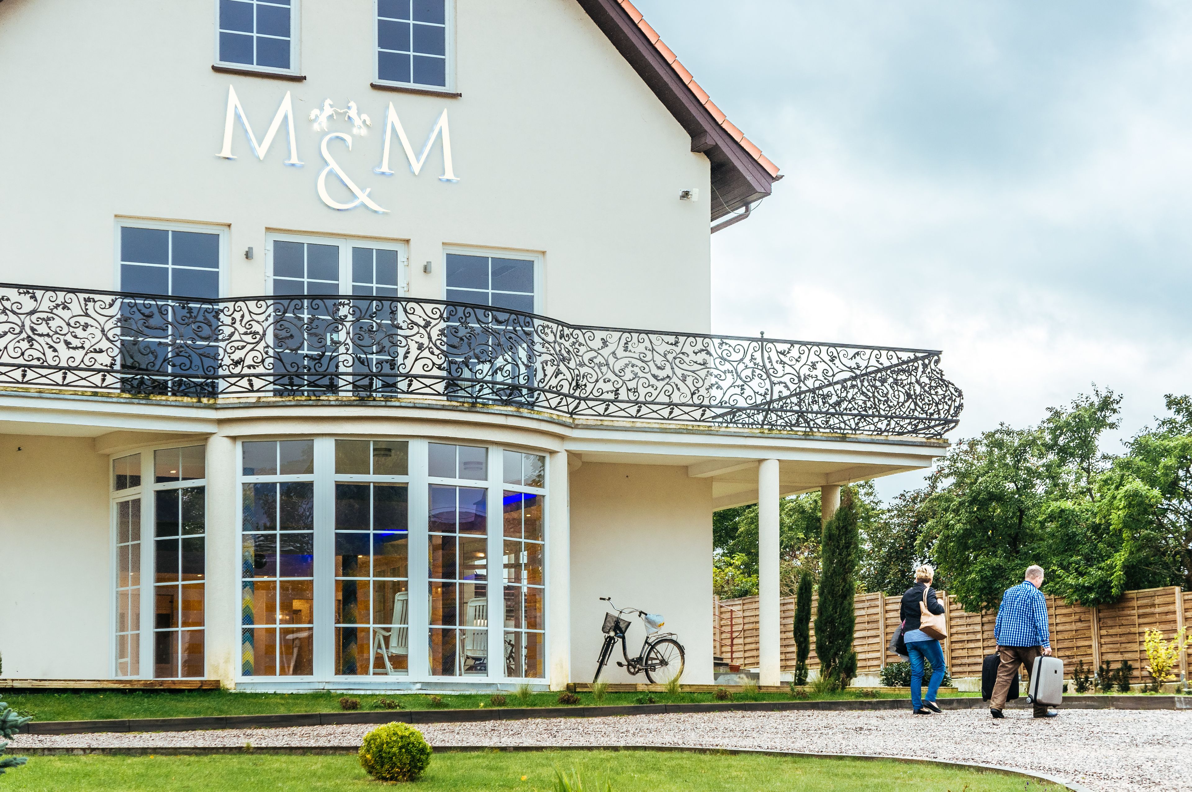 A building with a large sign reading "M&M" on the facade, a curved balcony with ornate metal railings, and a pathway leading to the entrance.