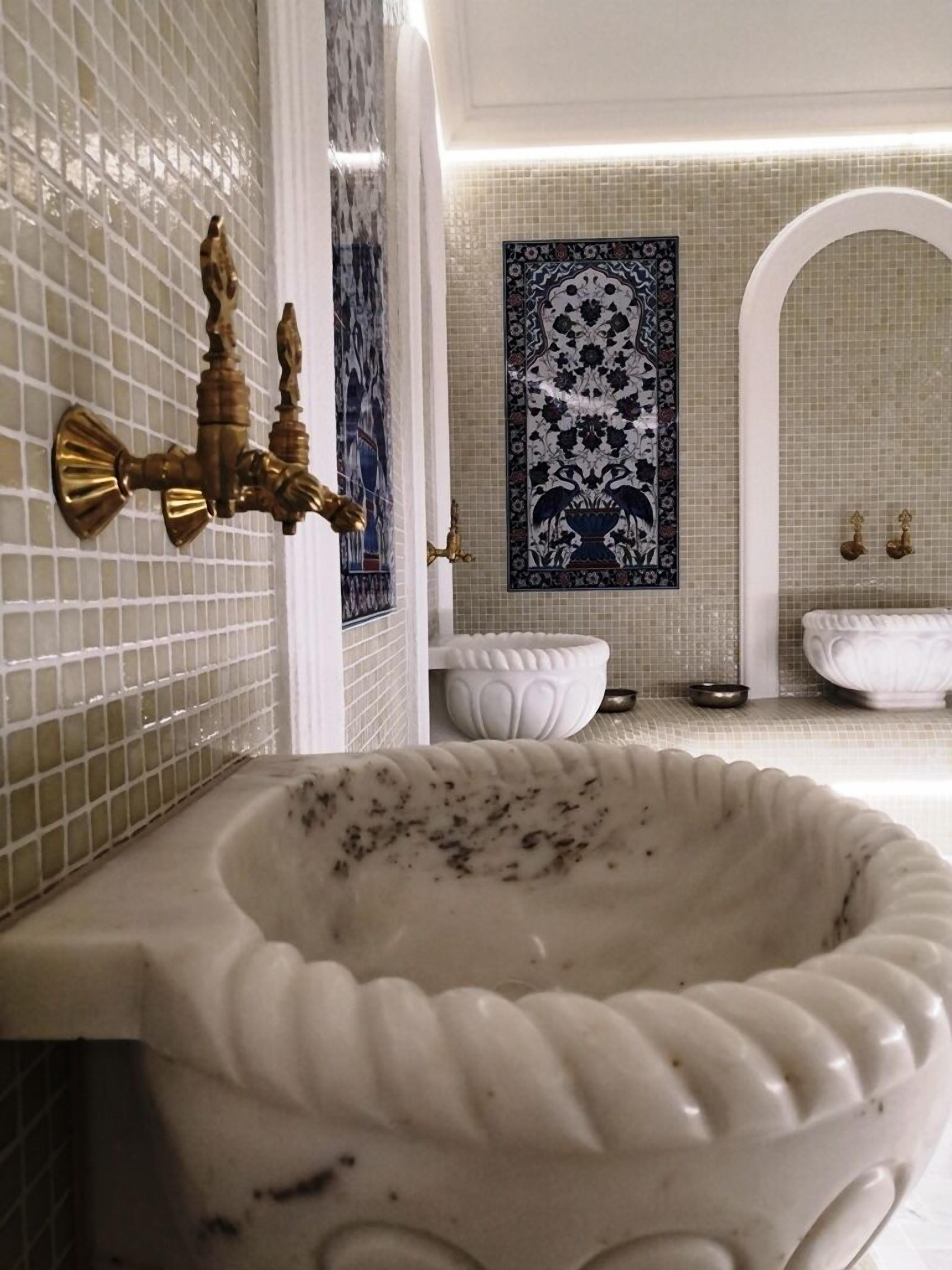 Un baño con un lavamanos grande y ornamentado, un grifo de bronce empotrado en la pared y un salpicadero decorado con azulejos.