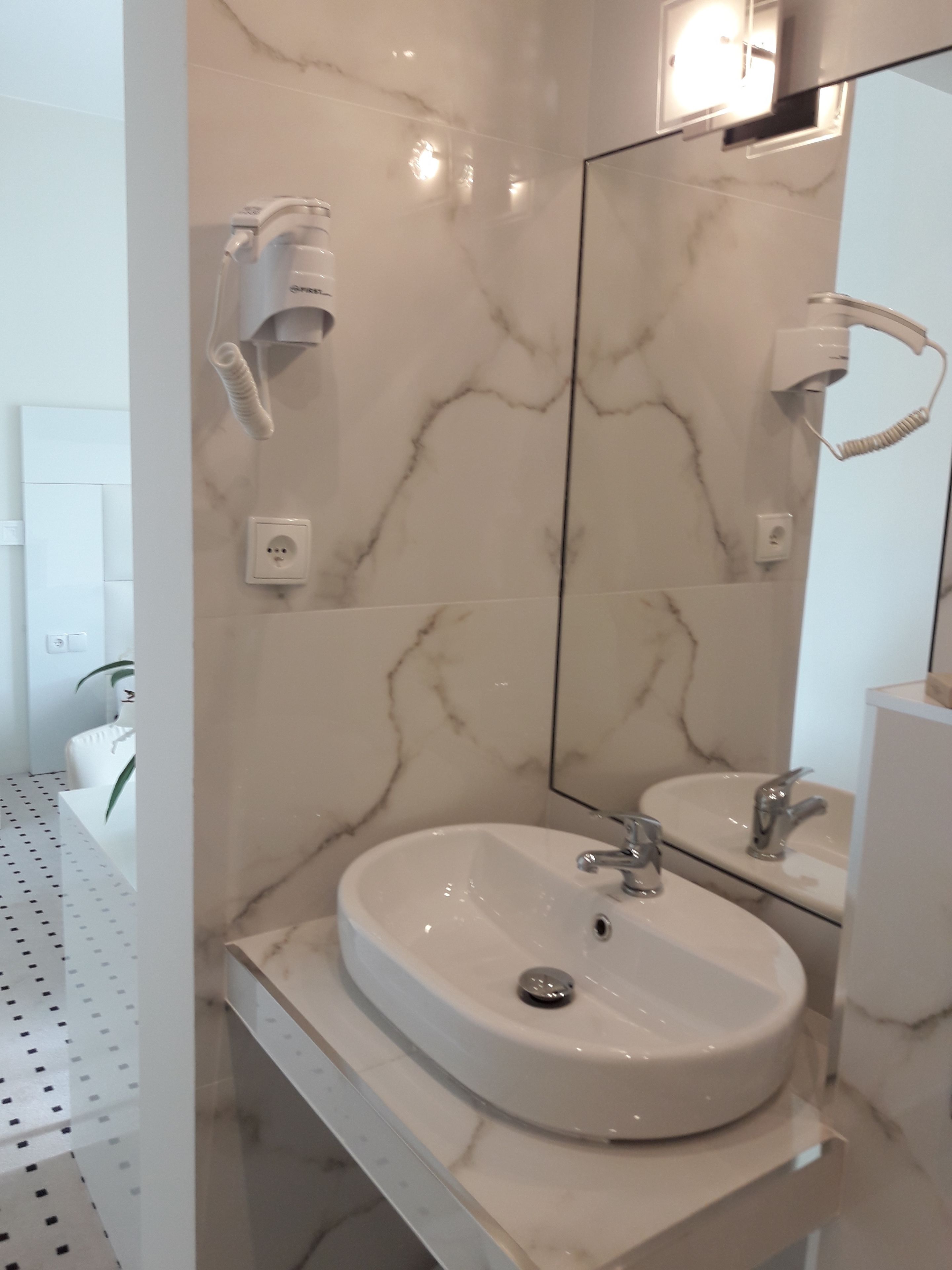 Un baño con pared de mármol, un lavamanos, un espejo y un secador de pelo fijado a la pared.