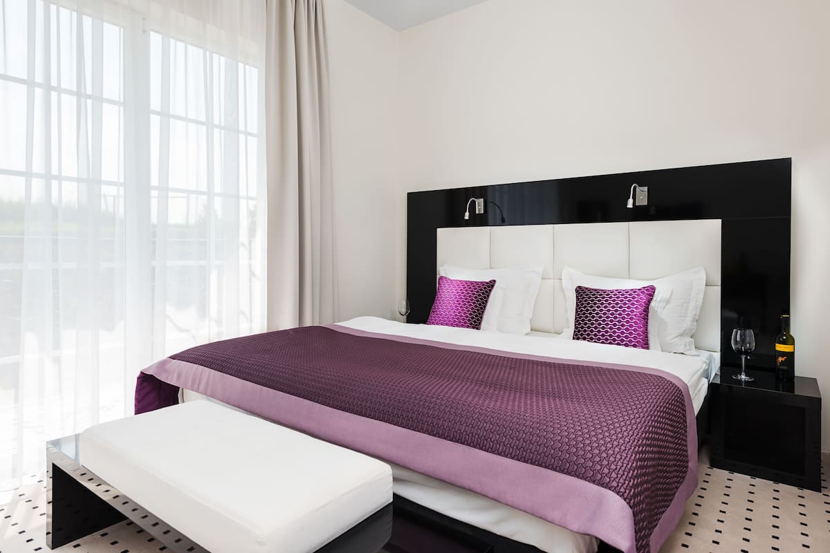 A modern hotel room with a large bed, a bedside table, and a window with sheer curtains.