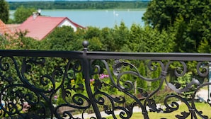 A balcony with ornate metal railings overlooking a lake with boats, surrounded by greenery and a red-roofed building. - Boutique Apartmenty M&M (Mikolajki)