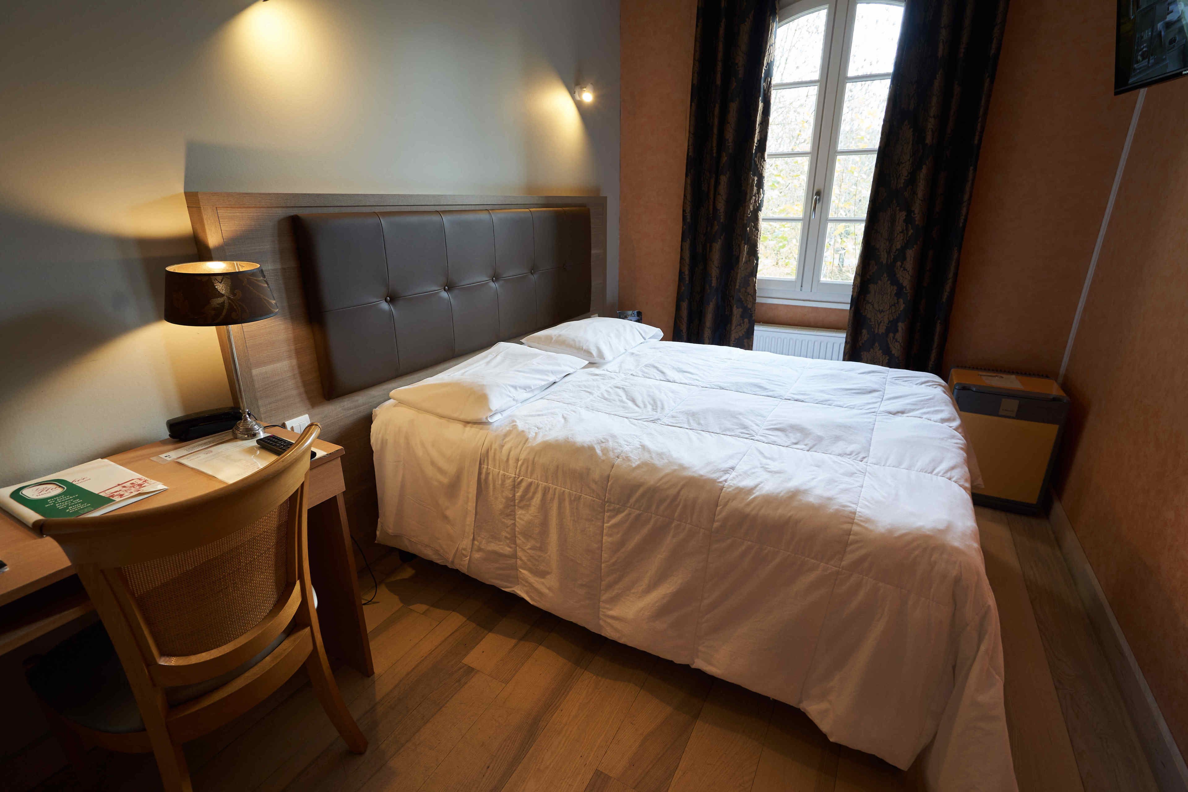 Een hotelkamer met een bed, een bureau, een stoel, een lamp en een raam met gordijnen.