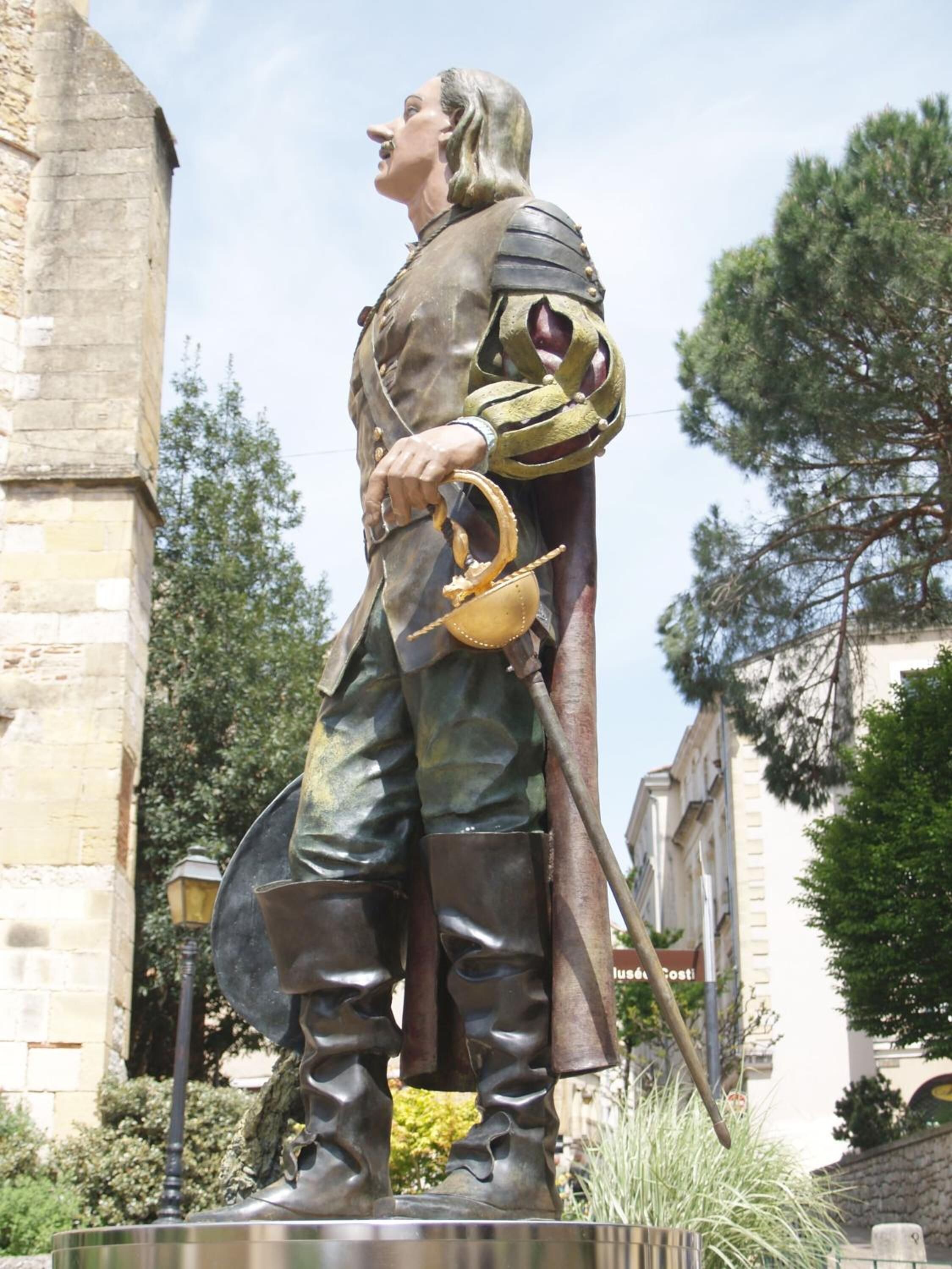 Una estatua de un personaje histórico con armadura, sosteniendo una espada y un escudo, sobre un pedestal en un entorno exterior con edificios y árboles de fondo.