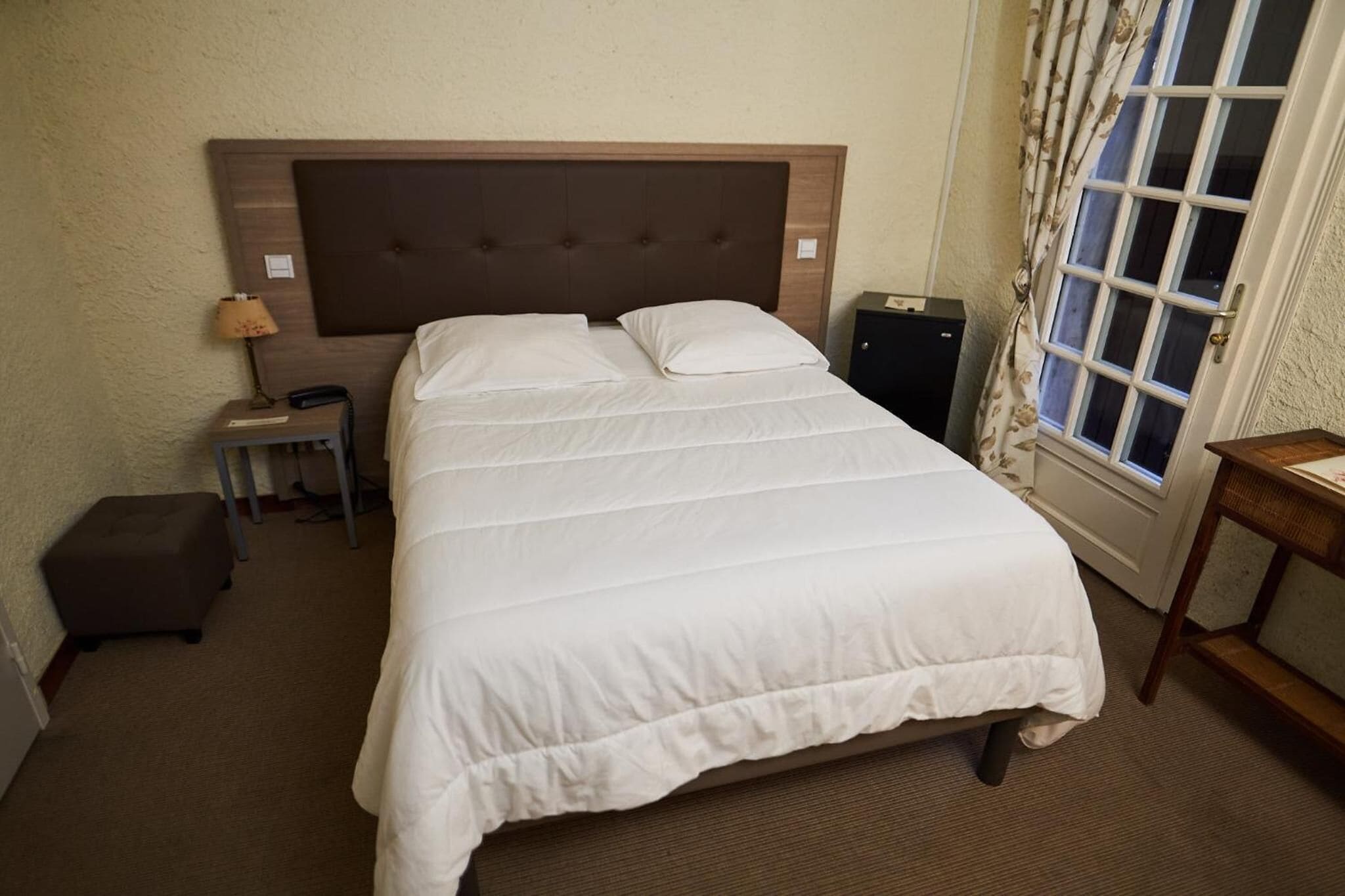 A hotel room with a large bed, a bedside table with a lamp, a small ottoman, and a window with curtains.