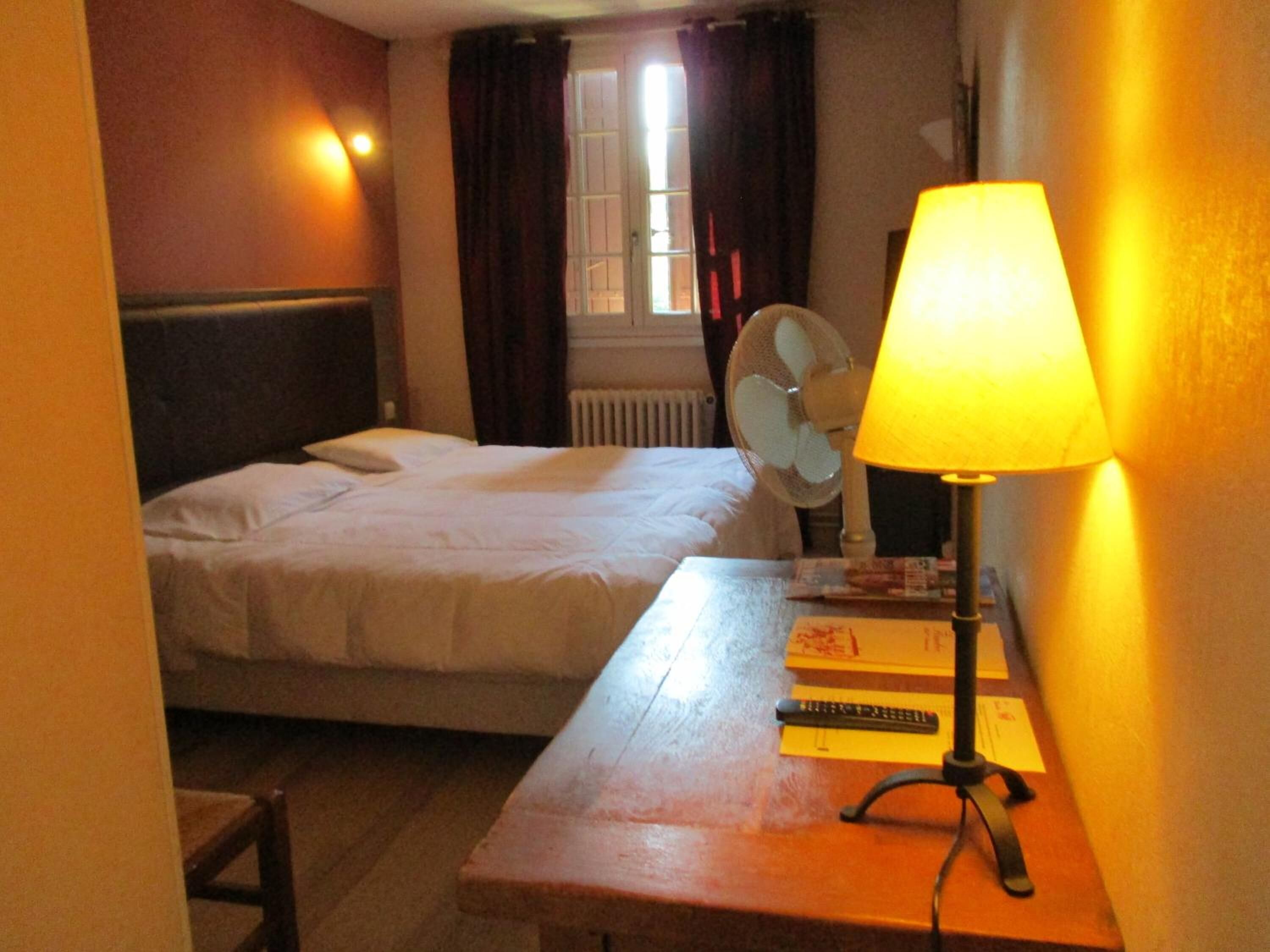 Une chambre d’hôtel avec un lit, un bureau avec une lampe, un ventilateur et une fenêtre avec des rideaux.