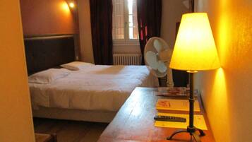 Une chambre d’hôtel avec un lit, un bureau avec une lampe, un ventilateur et une fenêtre avec des rideaux.