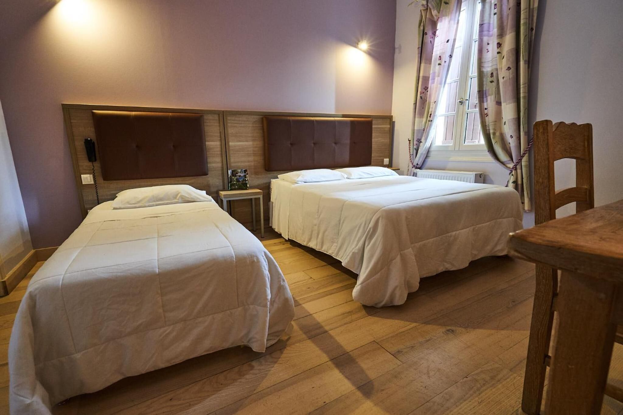 Habitación de hotel con dos camas individuales, cabecera de madera, mesita de noche con lámpara y ventana con cortinas.