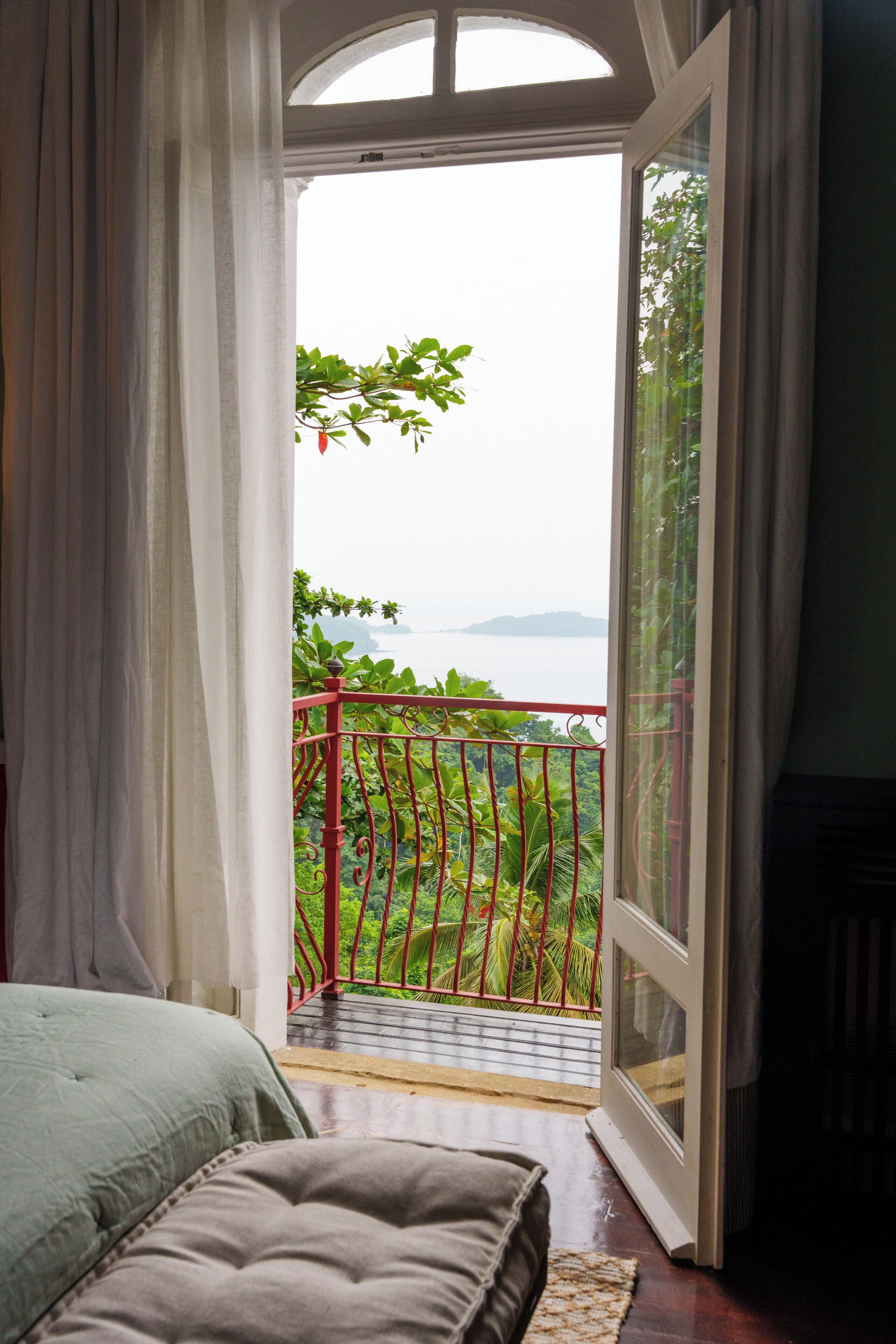 Un dormitorio con cama, ventana y vista a un balcón con vegetación y un cuerpo de agua.