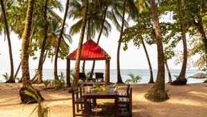 A beach setting with a wooden table and chairs, a red-roofed gazebo, and palm trees.