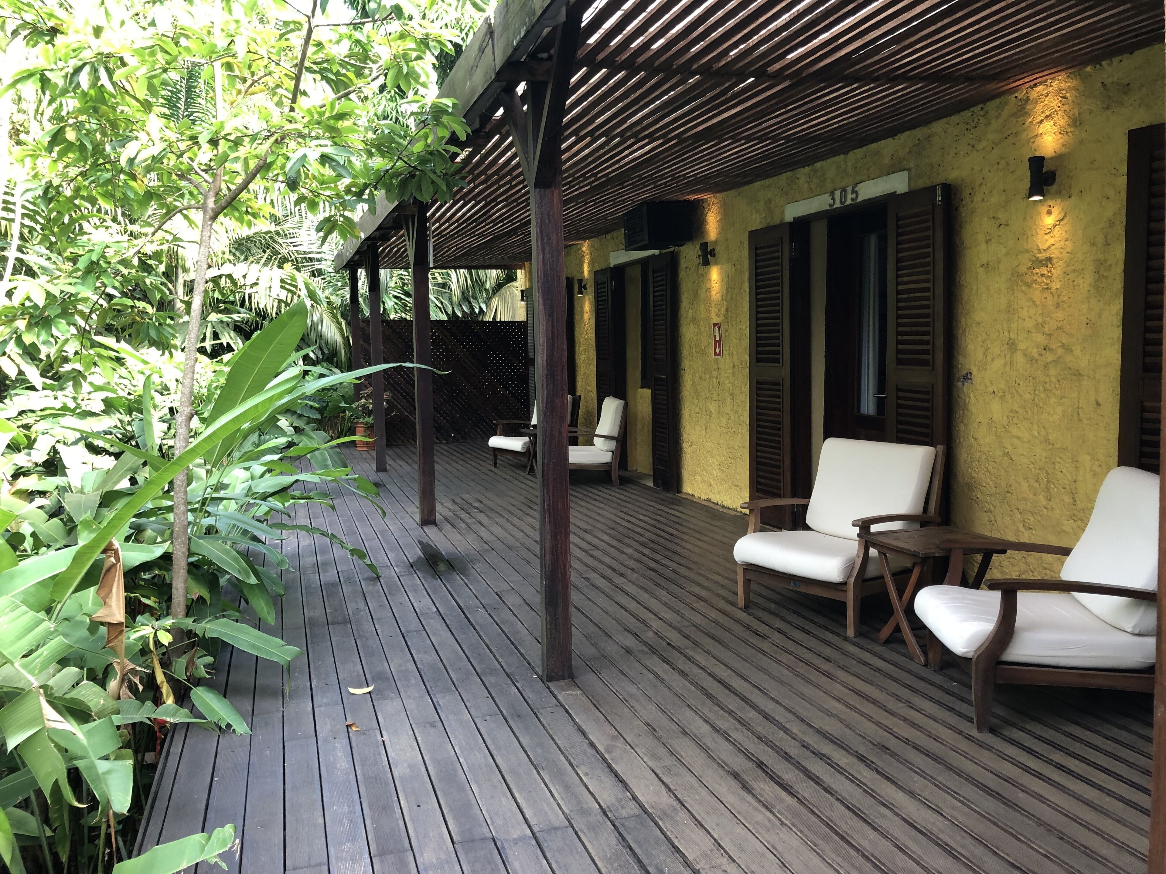 A covered outdoor area with wooden flooring, white cushioned chairs, and a yellow wall.