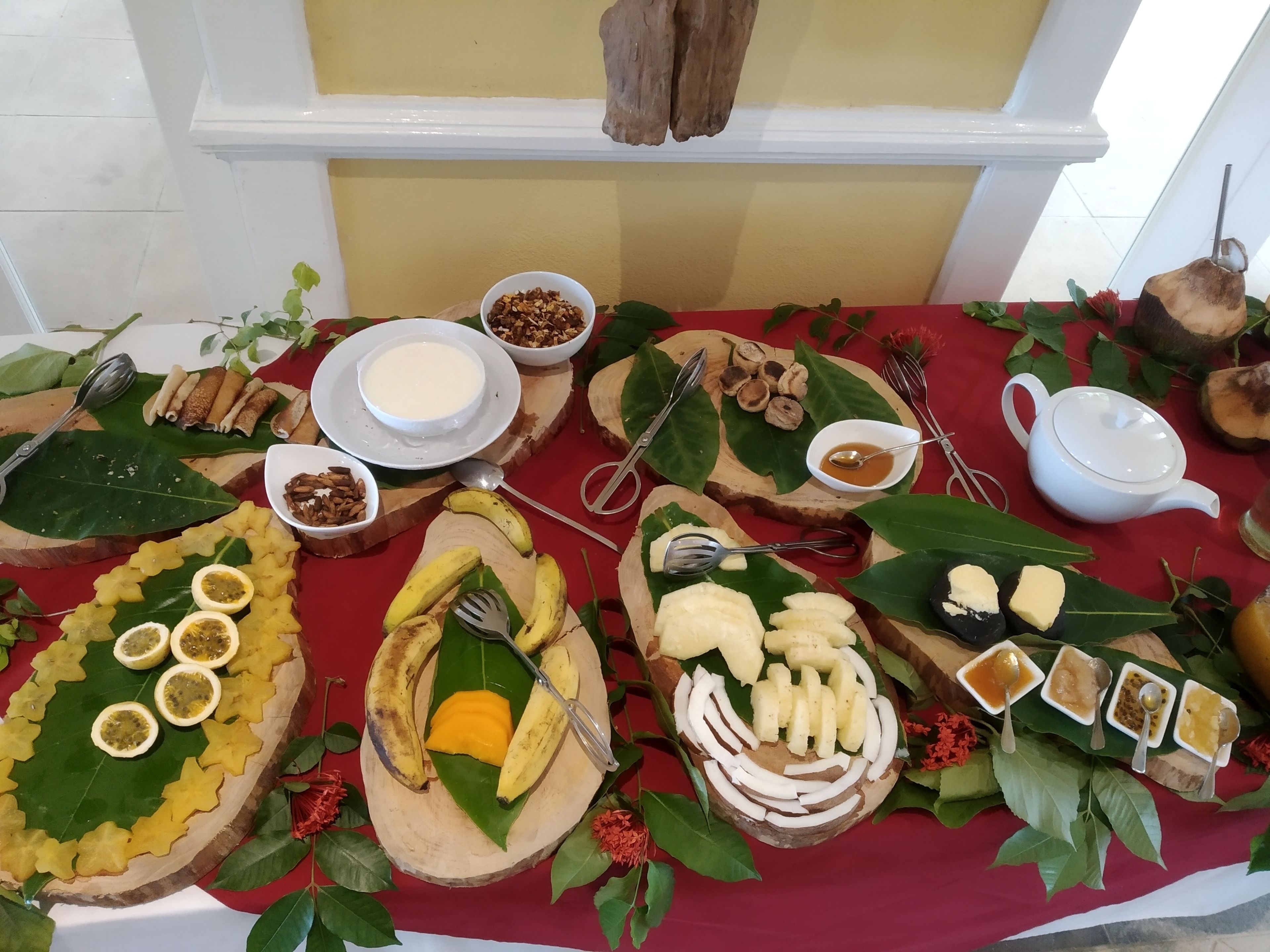 A table with various food items and utensils, including bananas, mango slices, and bowls of nuts and sauces.